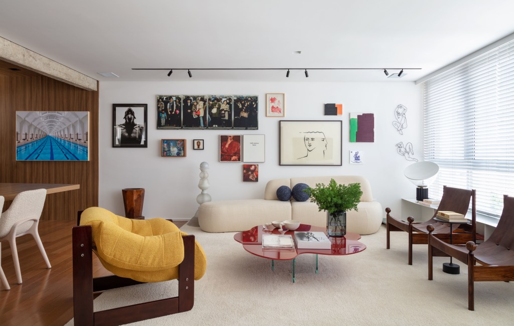 Sala de estar com tapete off white, sofá curvo, poltrona amarela, mesa de centro vermelha e quadros atrás do sofá.