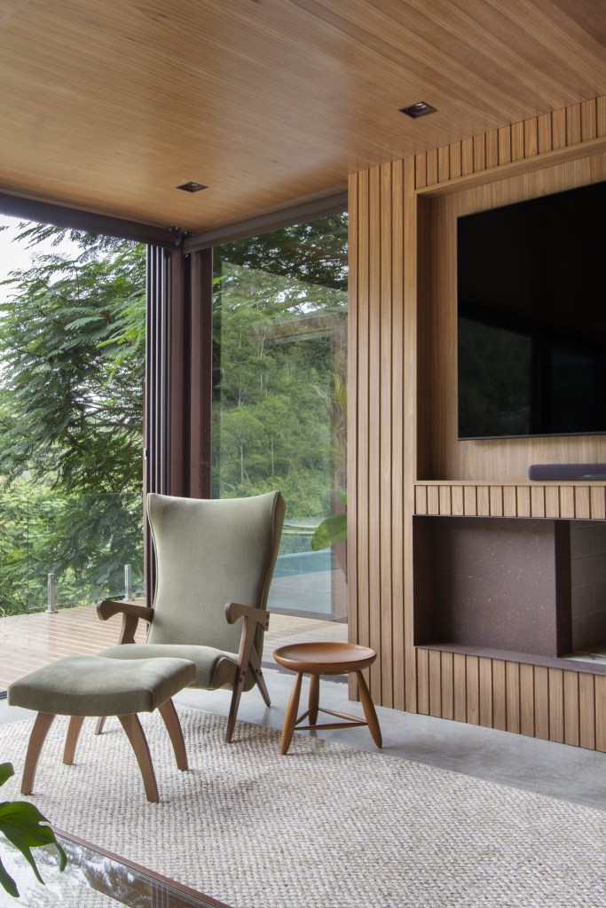 Sala com portas de vidro grandes com vista para serra, parede e teto revestido de madeira, poltrona verde e painel ripado para tv.