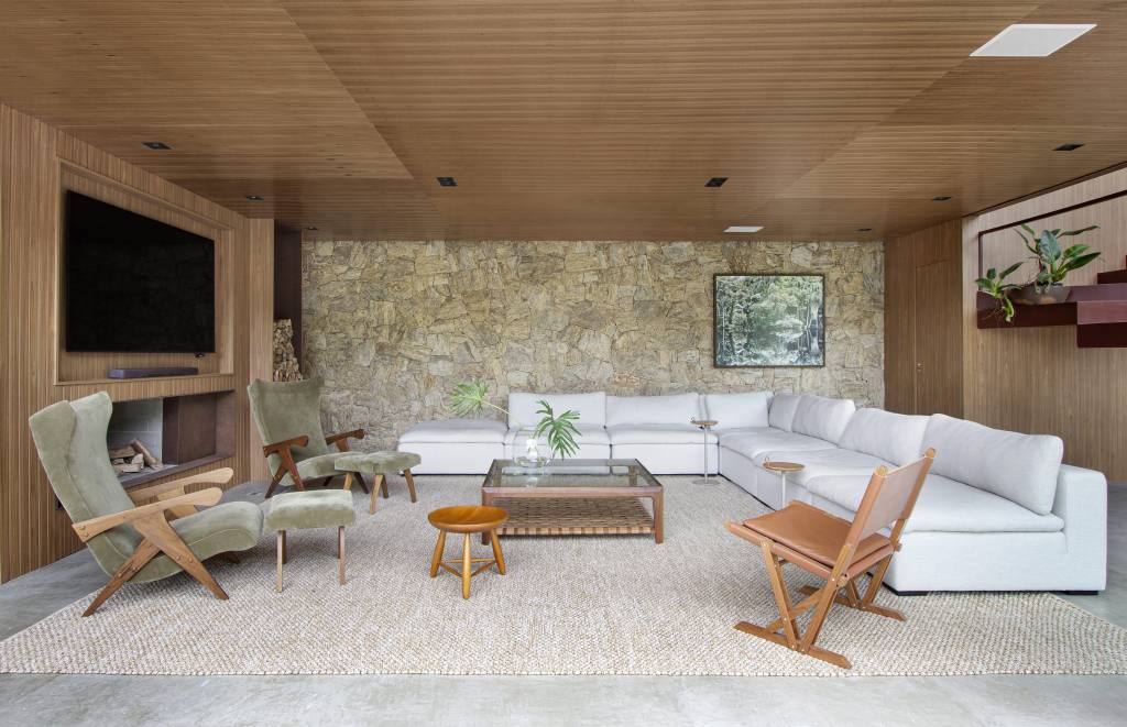 Sala de estar com parede ripada, teto de madeira e parede revestida de pedras; sala com sofá branco, tapete bege e mesa de centro de vidro.