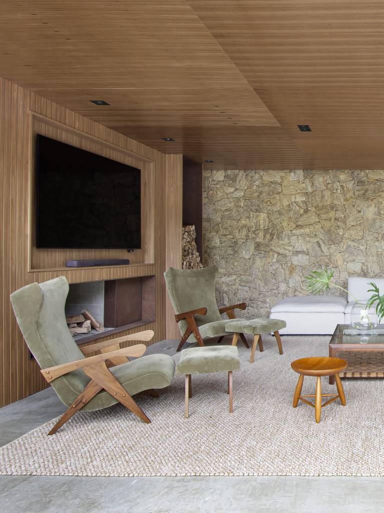 Sala com portas de vidro grandes com vista para serra, parede e teto revestido de madeira, sofá claro, mesa de centro, tapete bege, parede de pedras.