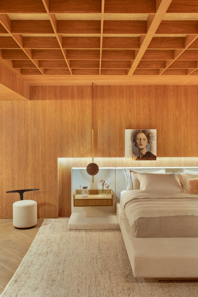 Quarto com cama de casal, com parede revestida de madeira, luminária lateral, mesa lateral.