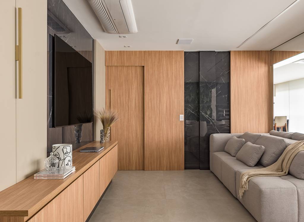 Paineis de porcelanato e jardim vertical revestem paredes em apê de 134 m². Projeto de Bianca Bahlis. Na foto, sala de tv com paines de porcelanato na parede, madeira e sofá cinza.