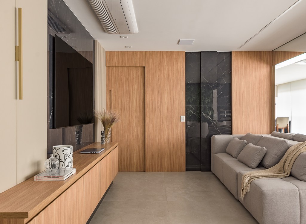 paineis-porcelanato-jardim-vertical-paredes-ape-134-m2-bianca-bahlis-credito-luiz-franco (2) Paineis de porcelanato e jardim vertical revestem paredes em apê de 134 m². Projeto de Bianca Bahlis. Na foto, sala de tv com paines de porcelanato na parede, madeira e sofá cinza.