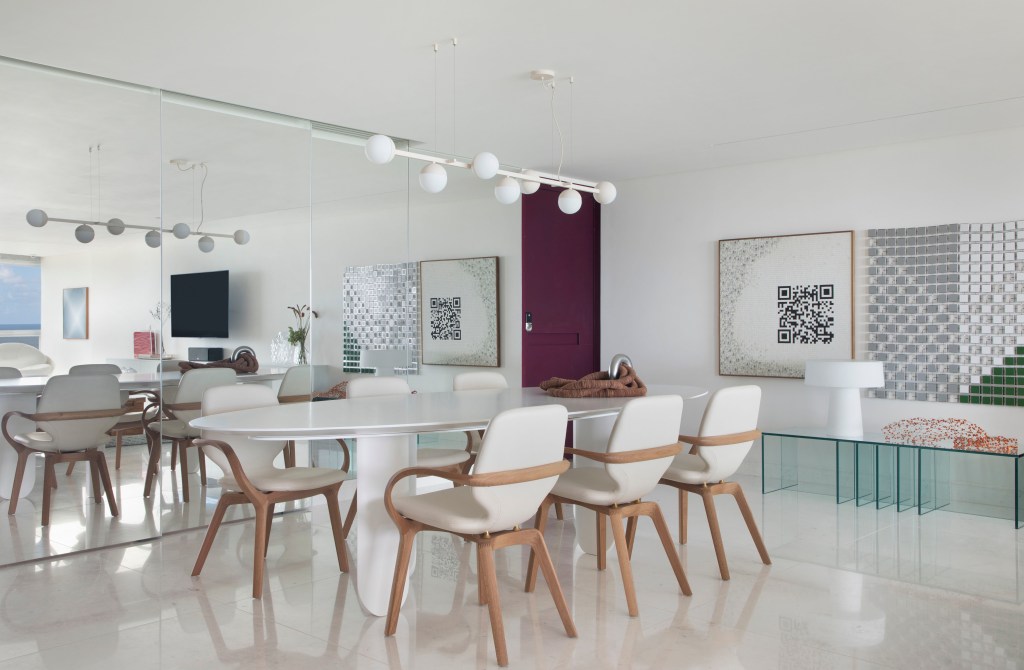Sala de jantar com piso de mármore, mesa branca, cadeiras estofadas brancas e luminária Ana Neute; parede espelhada ao fundo.