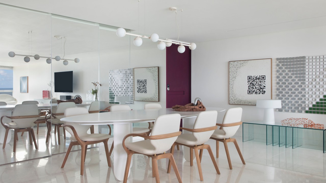 Sala de jantar com piso de mármore, mesa branca, cadeiras estofadas brancas e luminária Ana Neute; parede espelhada ao fundo.
