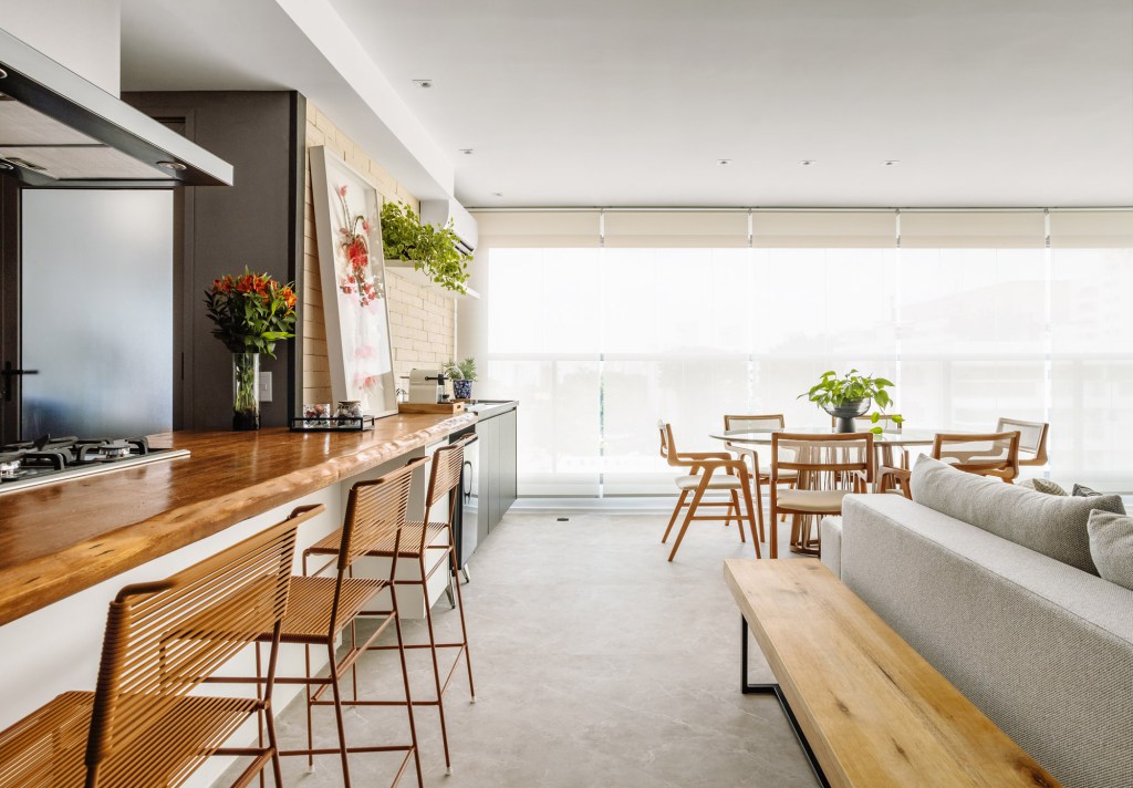 Sala integrada com varanda e cozinha; piso vinílico marmorizado, mesa redonda, bancada de madeira e sofá.