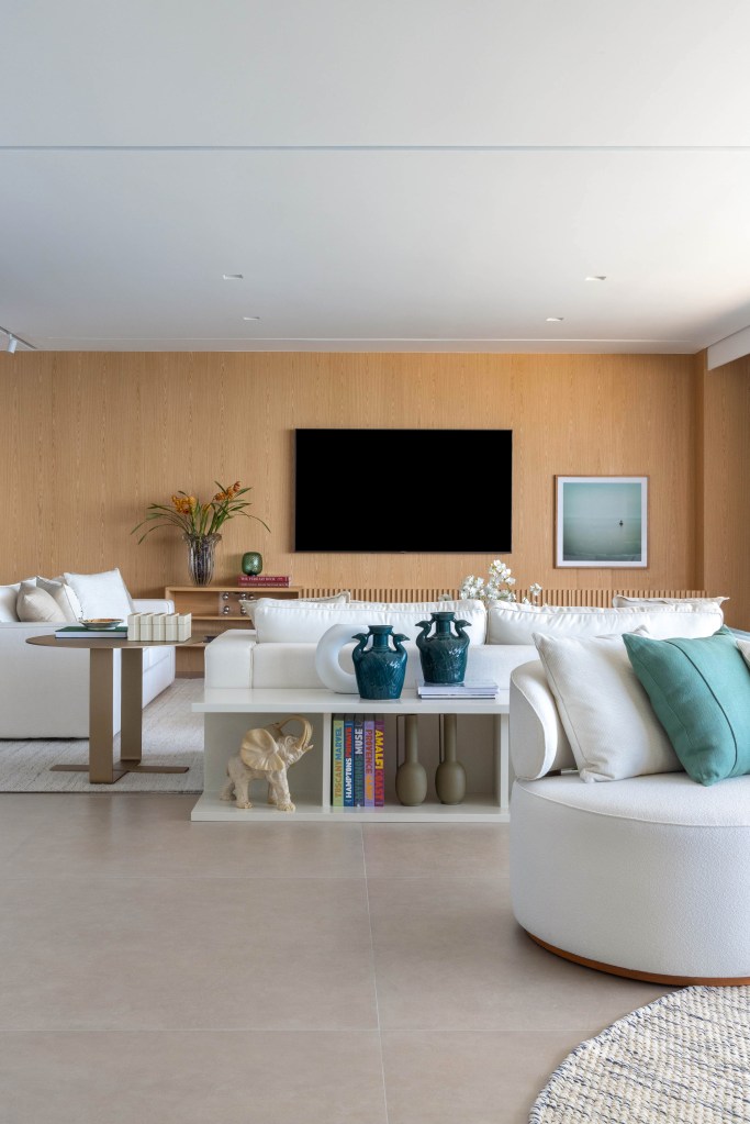 Sala de estar clara com piso de porcelanato, sofás brancos, tv e paredes revestidas de madeira.