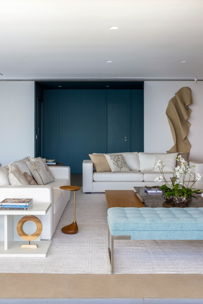 Sala de estar clara com dois sofás brancos, mesa de centro de madeira e pufe azul.