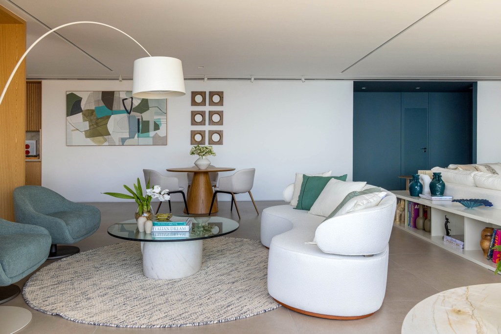 Sala de estar com piso de porcelanato, sofá curvo branco, luminária de piso e tapete felpudo redondo.