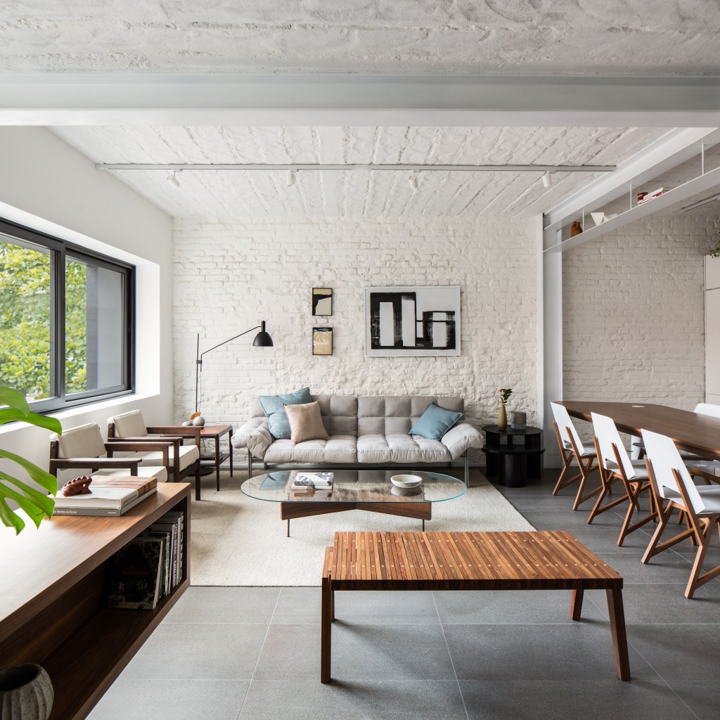 Sala de estar integrada com tapete claro, sofá cinza baixo e paredes com tijolos brancos expostos.