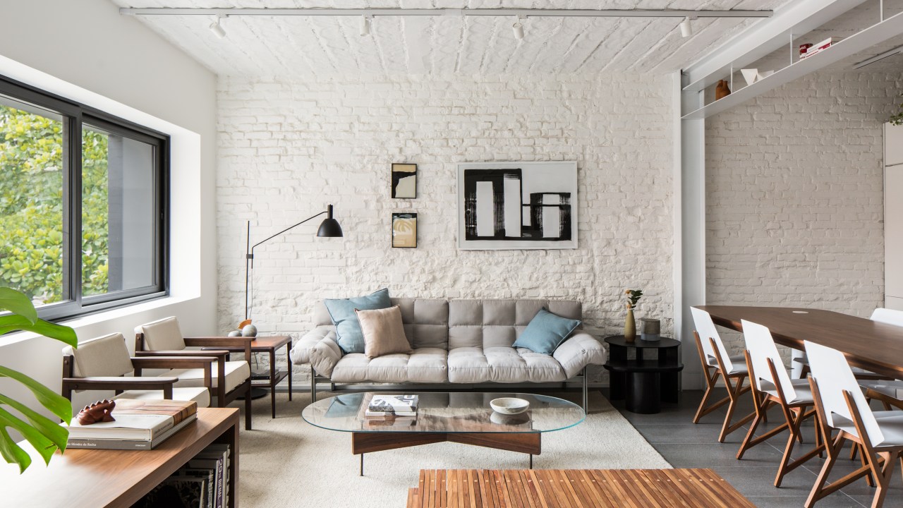 Sala de estar integrada com tapete claro, sofá cinza baixo e paredes com tijolos brancos expostos.