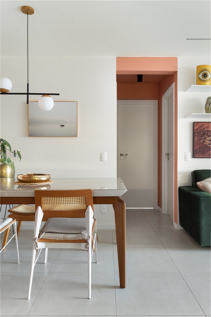Apartamento colorido de 65 m² é cheio de referências ao pop art. Projeto de Pistache Arquitetura. Na foto, sala de jantar com mesa de vidro e madeiral, corredor com parede rosa e sofá.