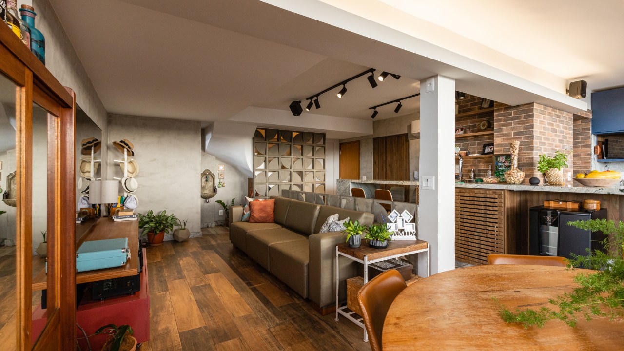 Sala de estar integrada com piso de madeira, mesa redonda e parede de tijolinhos na cozinha.