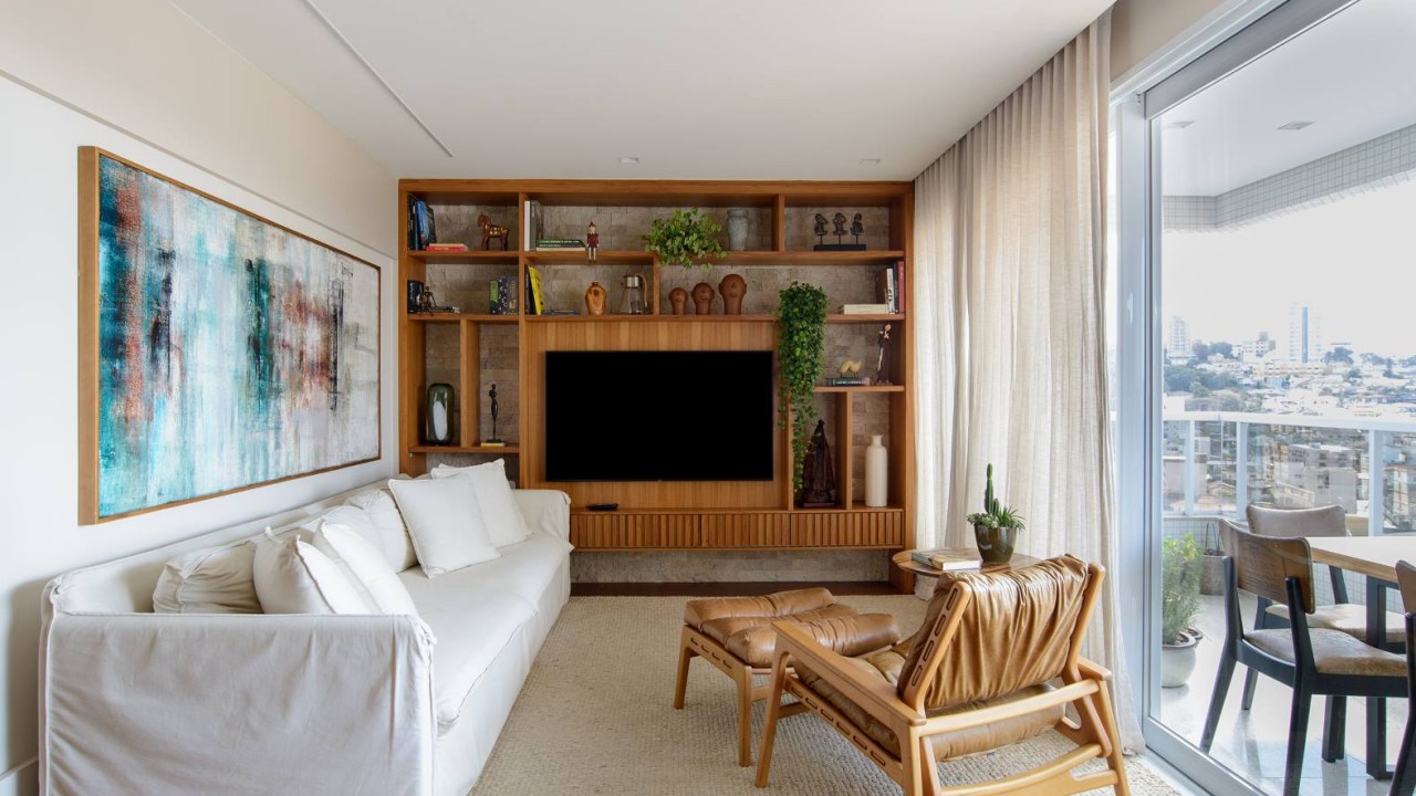 Apê de 140 m² feito para receber une madeira e mármore no décor. Projeto de Letícia Nannetti. Na foto, sala de estar com varanda, estante e parede de pedra.