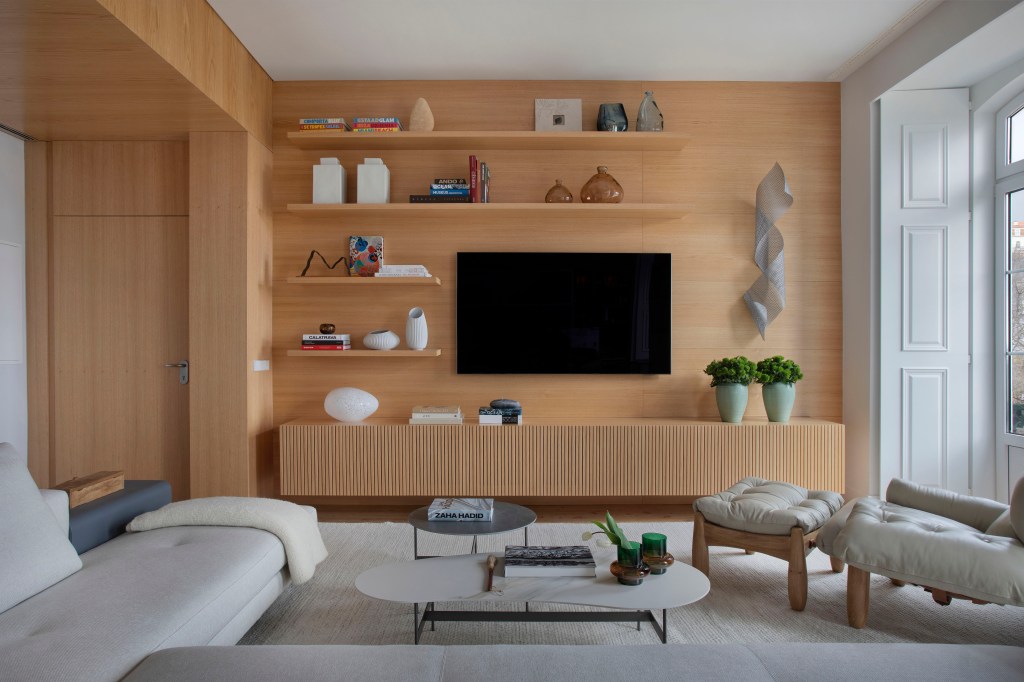 Sala de estar; sala de tv com parede revestida de madeira, rack de madeira, prateleiras, tv, tapete branco, sofá branco e poltrona mole.