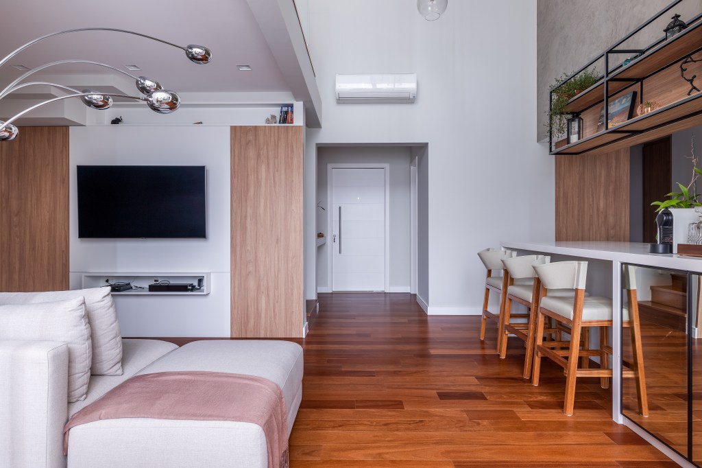 Sala de tv com piso de madeira, luminária de piso e sofá branco. Cozinha integrada.