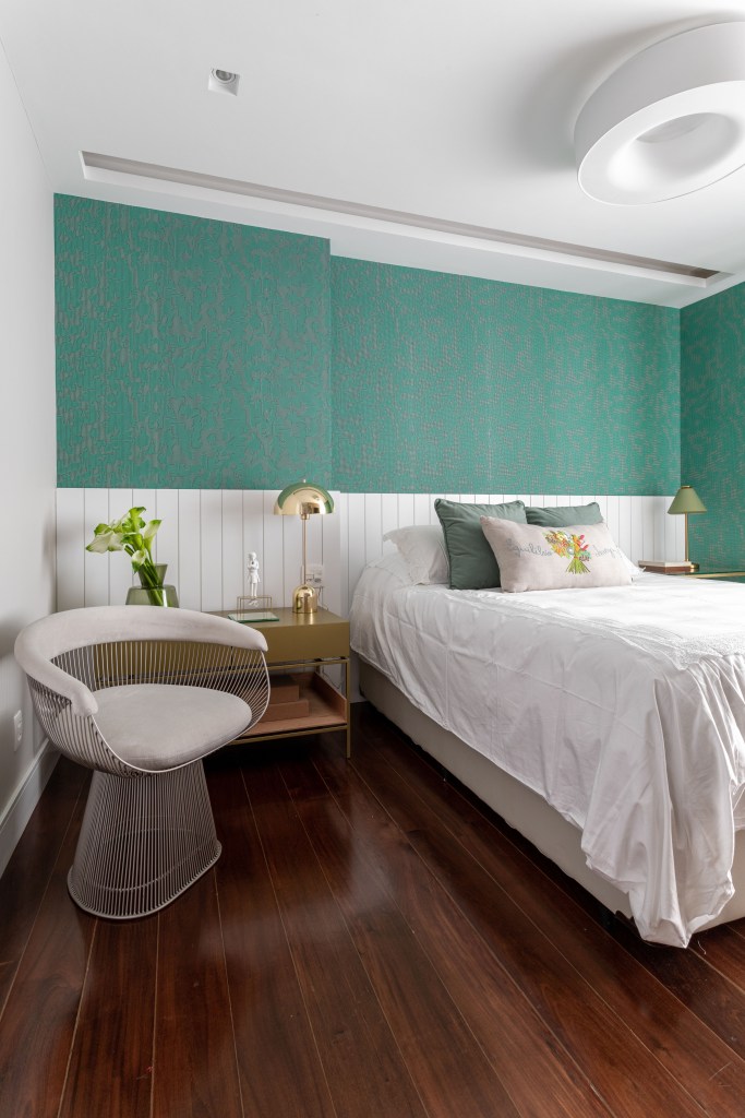 Quarto com cama de casal. cabeceira de lambris brancos e papel de parede com estampa azul esverdeada.