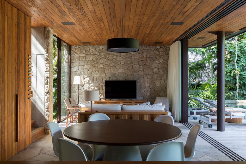 Casa de 153 m² se camufla em meio à natureza do litoral paulista. Projeto de Daniel Fromer, interiores de Julyana Bortolotto. Na foto, sala de tv com parede de pedra e mesa de jantar redonda.