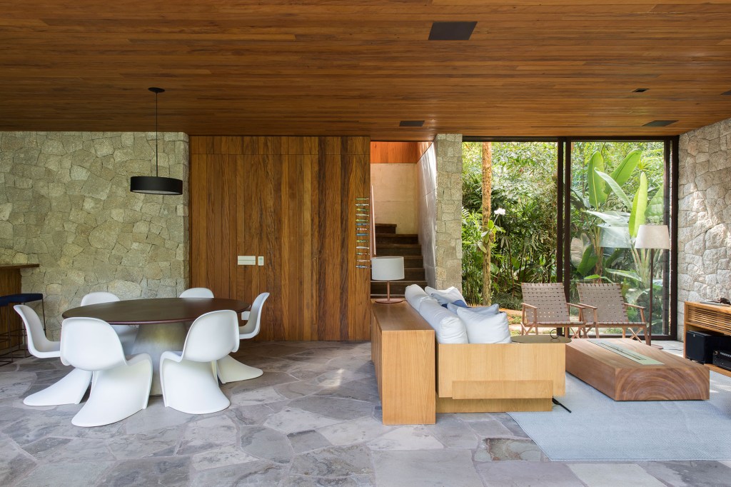 Casa de 153 m² se camufla em meio à natureza do litoral paulista. Projeto de Daniel Fromer, interiores de Julyana Bortolotto. Na foto, sala de estar com parede de vidro e escada.