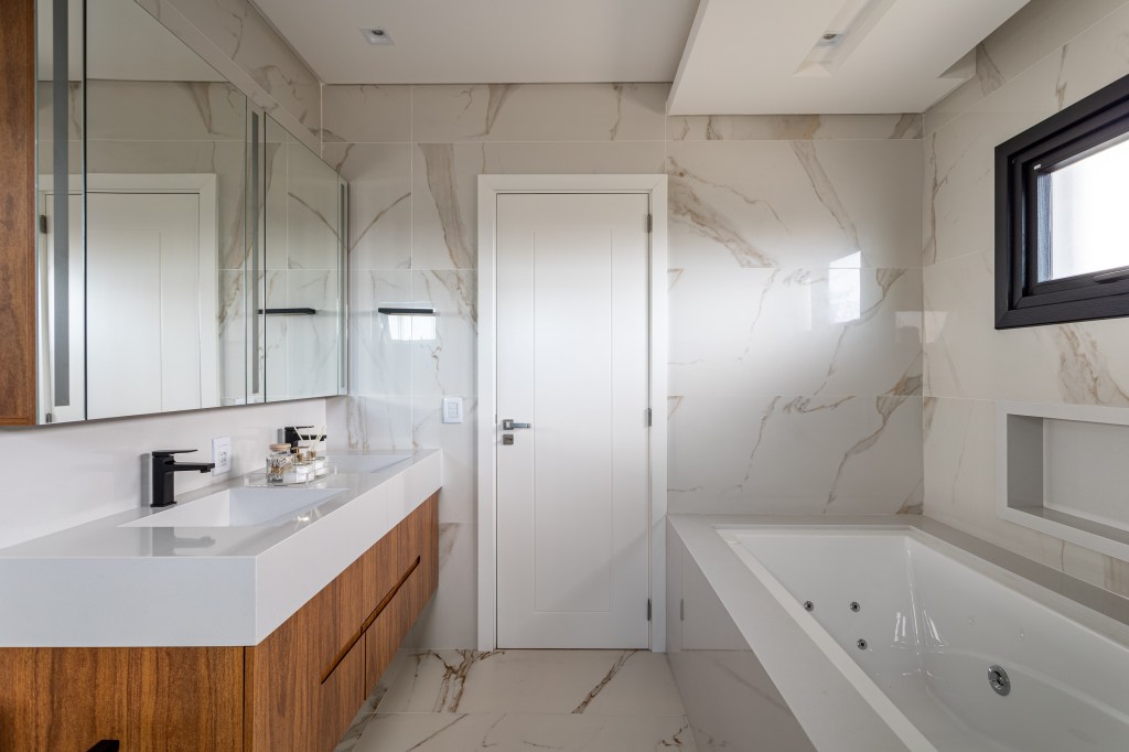 Banheiro com duas cubas esculpidas brancas, armário em madeira, banheira e revestimento marmorizado.