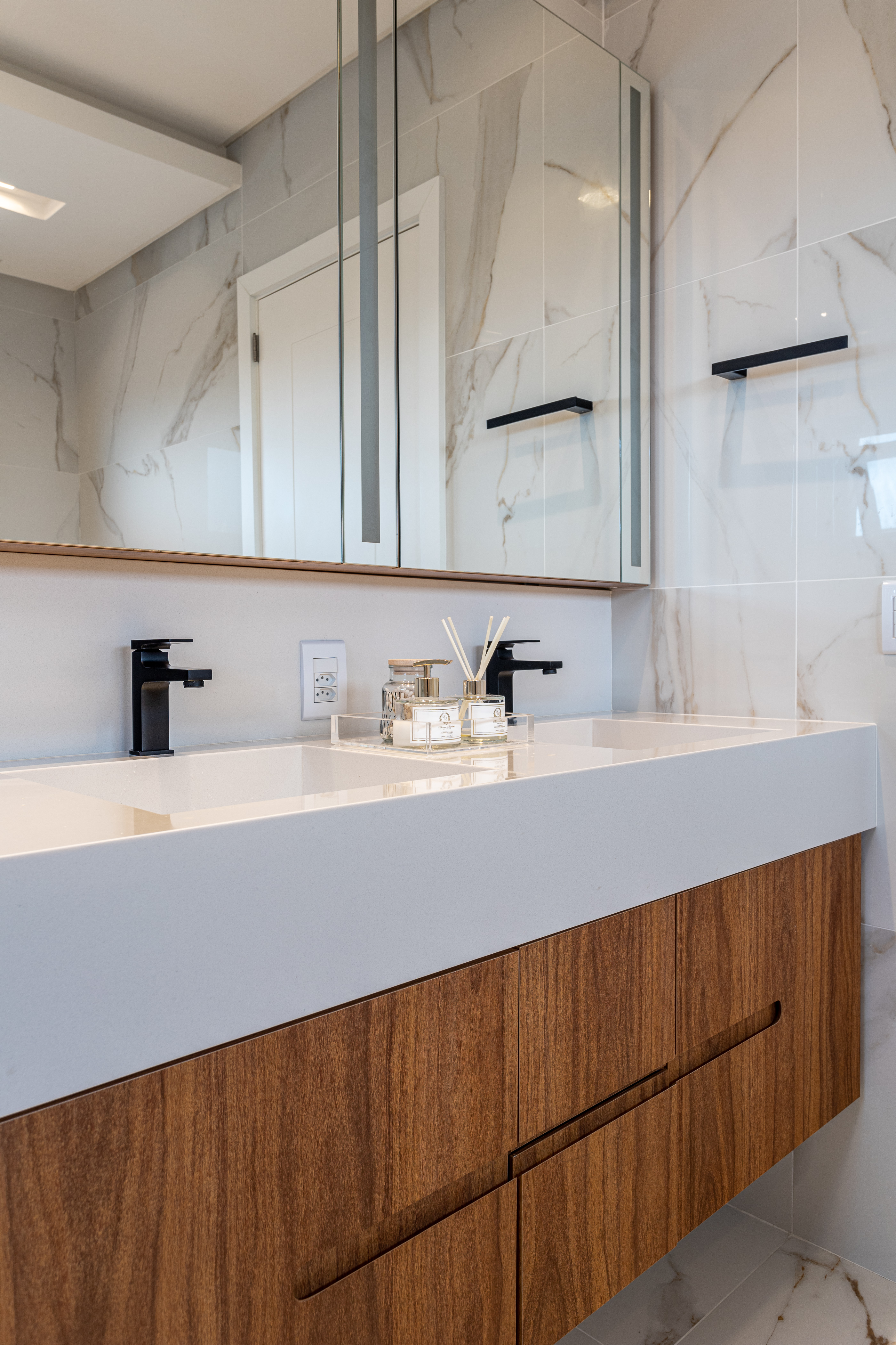 Banheiro com duas cubas esculpidas brancas, armário em madeira e revestimento marmorizado.