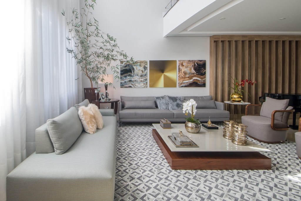 Casa de 500 m² possui detalhes dourados em todos os ambientes. Projeto de Ana Cano Milman. Na foto, sala de estar com sofá cinza, piano, tapete e mezanino.