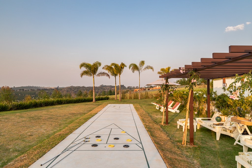 Casa de campo térrea tem vista para o laranjal e espaço para shuffleboard. Projeto de Gilda Meirelles. Na foto, varanda com gramado e campo de shuffleboard,