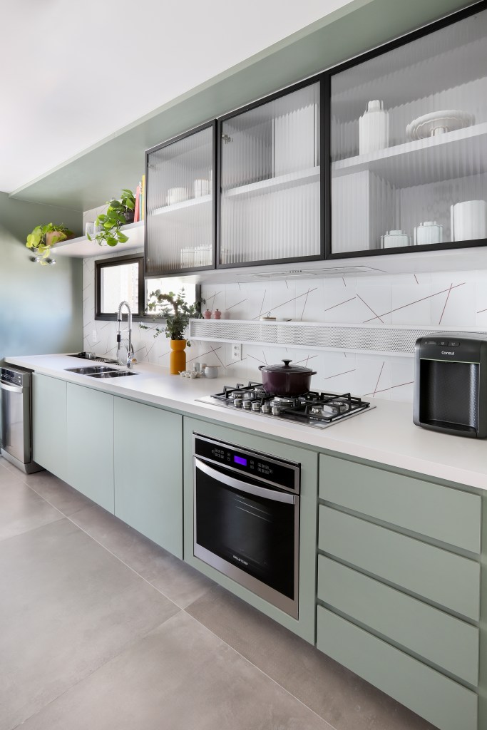 Cozinha integrada com piso de porcelanato cinza e marcenaria verde clara com armários com porta de vidro,