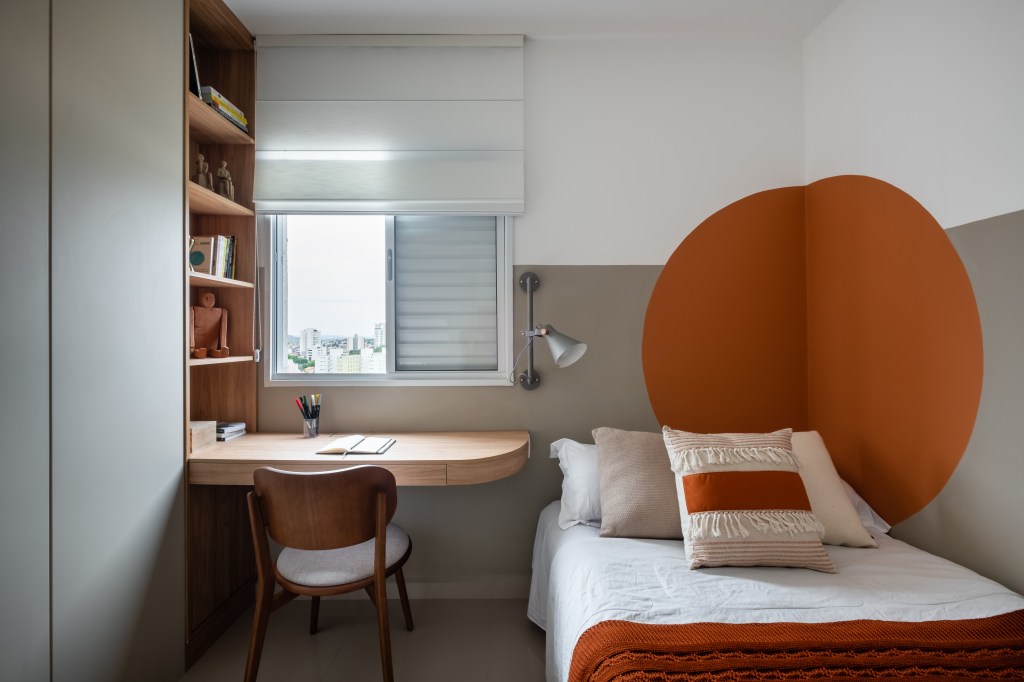 Quarto com meia parede cinza, círculo laranja e home office pequeno com bancada de madeira.