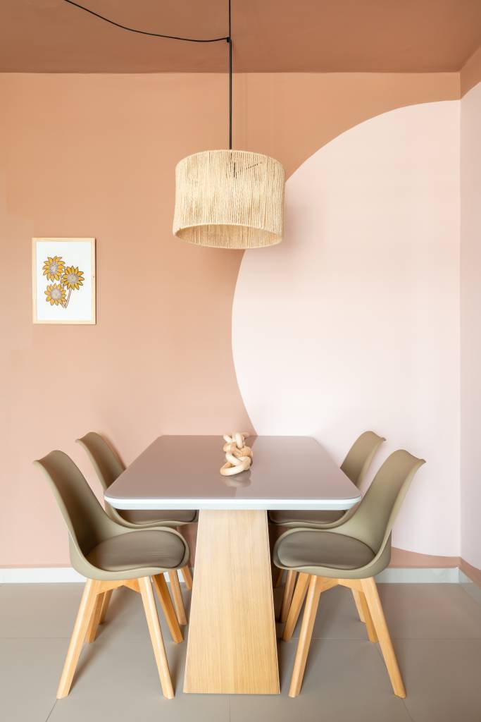 Sala de jantar pequena com mesa retangular e cadeiras taupe; círculo rosa claro na parede e luminária de palhinha.