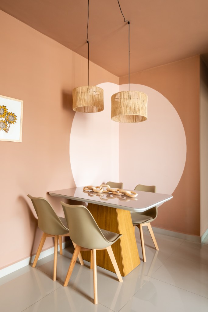 Sala de jantar pequena com mesa retangular e cadeiras taupe; círculo rosa claro na parede e luminária de palhinha.