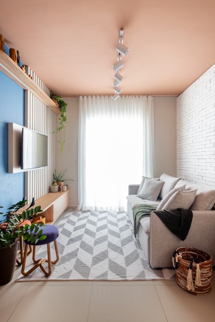 Sala de estar; sala de tv com parede azul, teto rosa, tapete com estampa geométrica preta e branca e sofá branco.