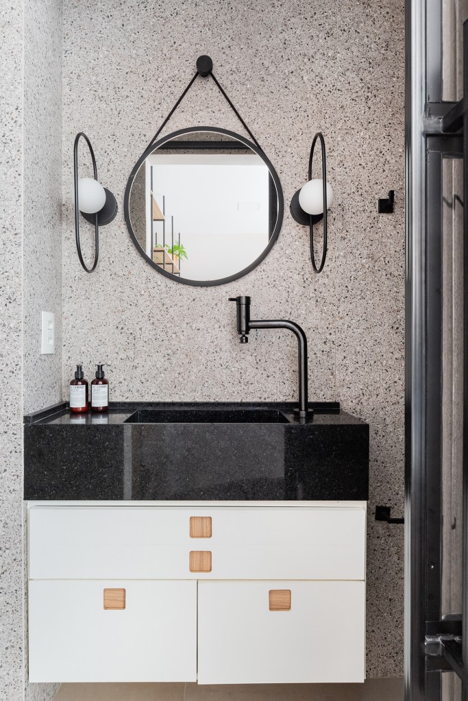 Estante de serralheria separa quarto e cozinha neste apê de 23 m². Projeto do Estúdio Taco. Na foto, banheiro com box de vidro canelado, metais pretos e espelho redondo.