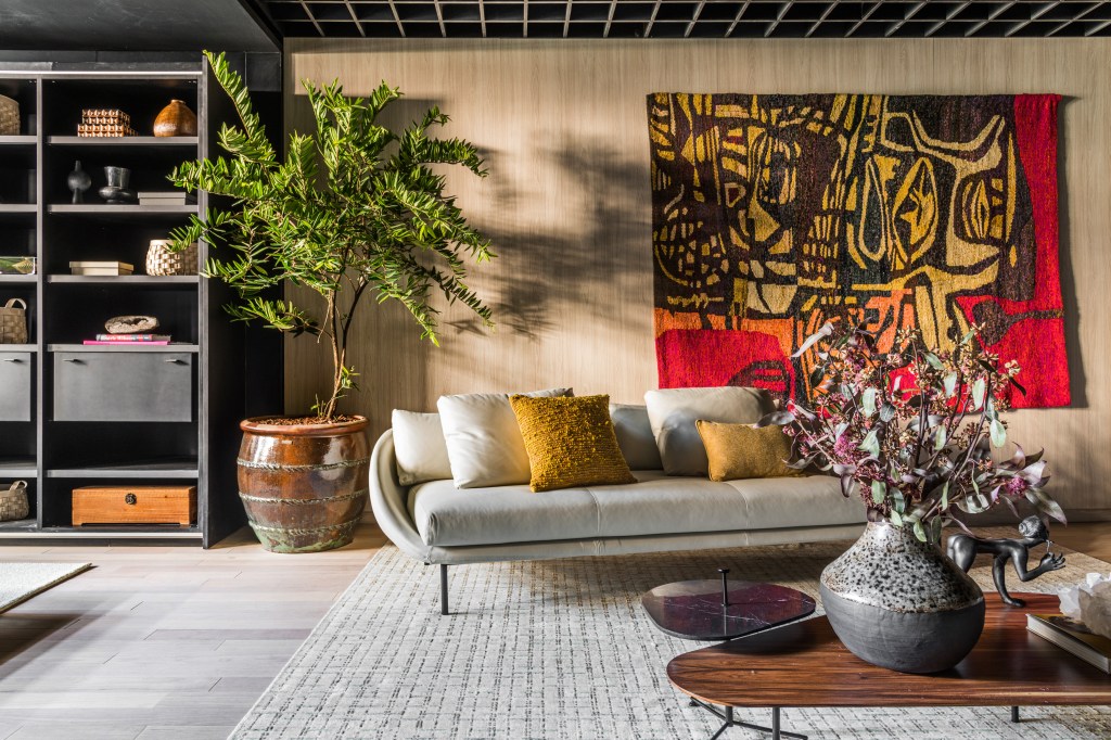 Estúdio de 66 m² tem jabuticabeira na bancada da cozinha e homenagem ao hipismo. Projeto de Erica Salgueiro para a CASACOR São Paulo 2023. Na foto, sala com sofá, tapete, estante e vaso.