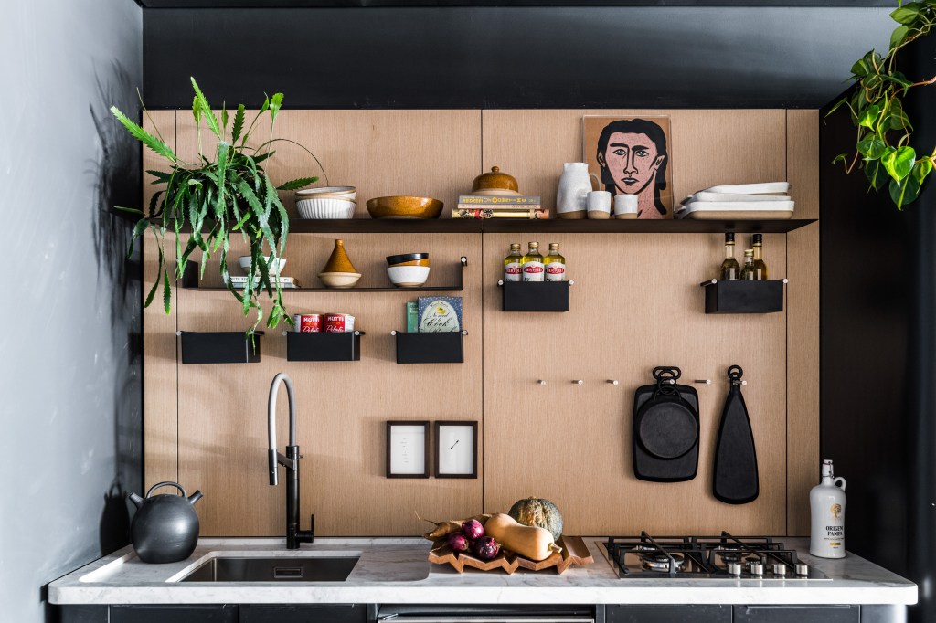Estúdio de 66 m² tem jabuticabeira na bancada da cozinha e homenagem ao hipismo. Projeto de Erica Salgueiro para a CASACOR São Paulo 2023. Na foto, cozinha com parede amadeirada, prateleiras e quadros.