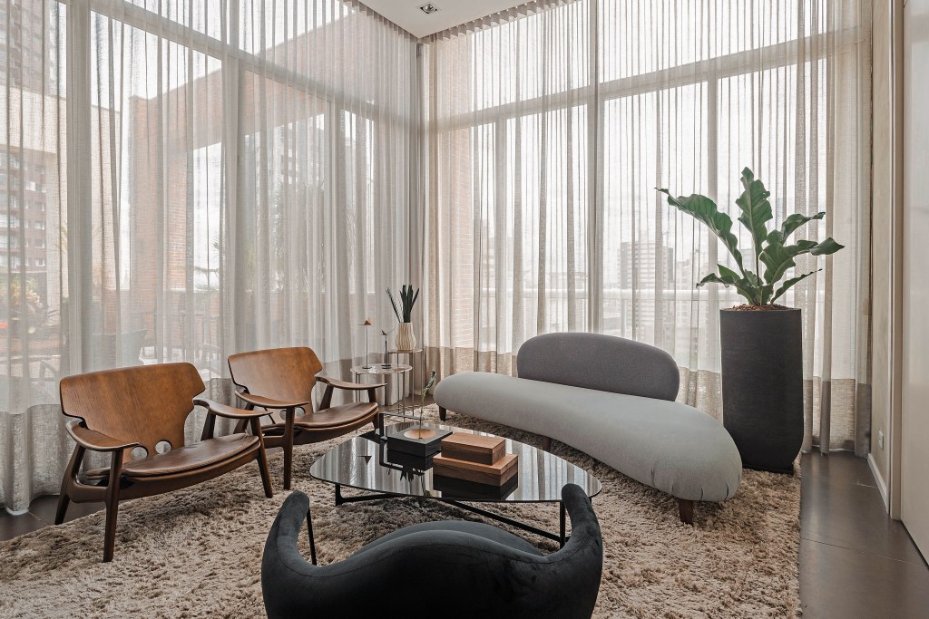 Grandes áreas envidraçadas marcam este dúplex de 220 m² em Curitiba. Projeto do Estudio Elmor. Na foto, sala de estar com janelas grandes e cortinas. Poltrona, sofá e tapete.