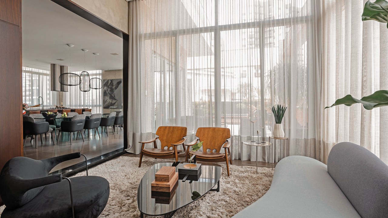 Grandes áreas envidraçadas marcam este dúplex de 220 m² em Curitiba. Projeto do Estudio Elmor. Na foto, sala de estar com janelas grandes e cortinas. Poltrona, sofá e tapete.
