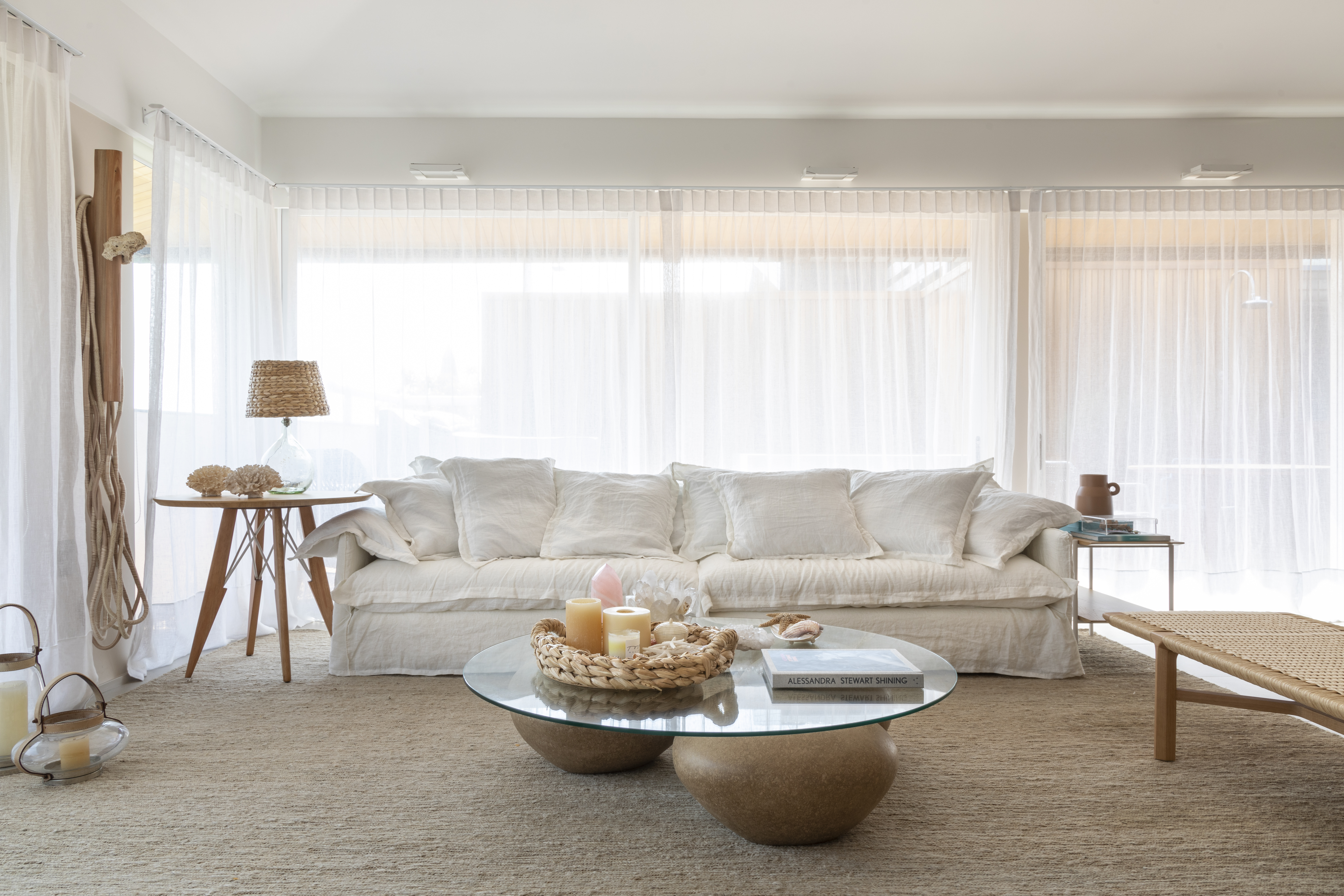 Sala de estar com tapete bege, sofá branco, cortina branca e mesa de centro com tampo de vidro.