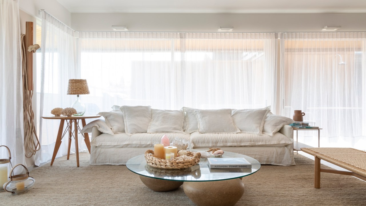 Sala de estar com tapete bege, sofá branco, cortina branca e mesa de centro com tampo de vidro.
