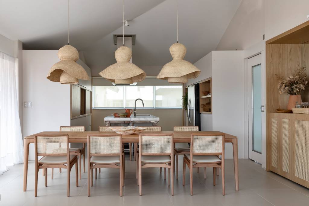 materiais-naturais-clima-praia-ape-alessandra-ambrosio-juliana-pippi-08 Sala de jantar integrada com cozinha com mesa de madeira e luminárias de palhinha.