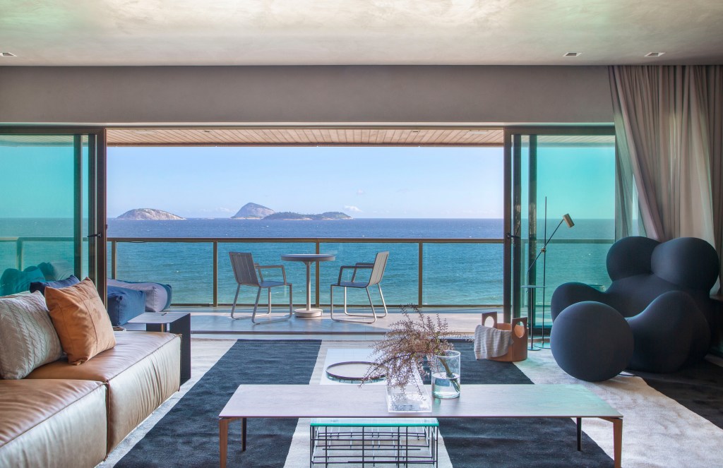 Sala com tapete azul e poltrona Up; varanda com vista para o mar.