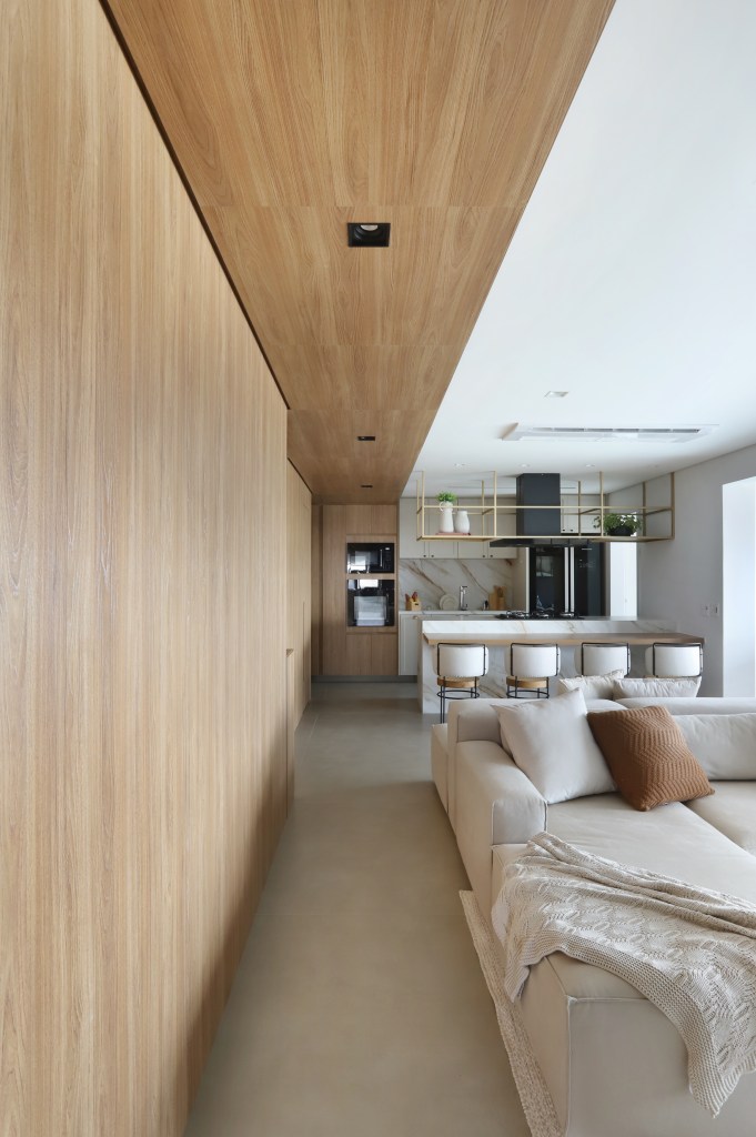 Painel de madeira percorre parede e teto da área social do apê de 120 m². Projeto Fantato Nitoli Arquitetura. Na foto, sala de estar com painel de madeira no teto, cozinha americana e sofá ilha.