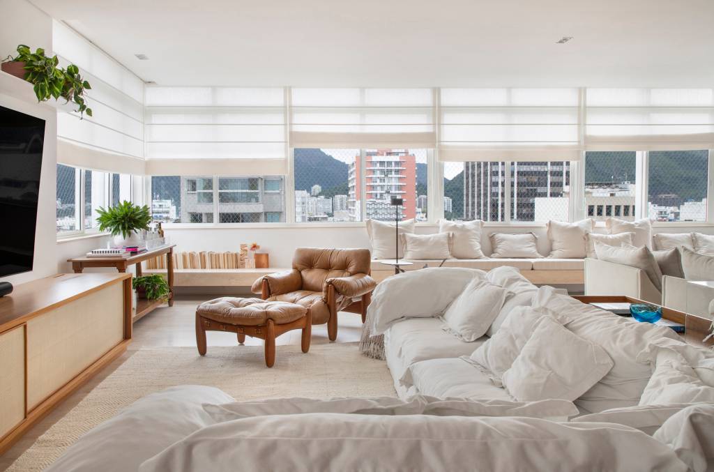 Sala de estar branca com janelas grandes, sofá branco e poltrona mole.