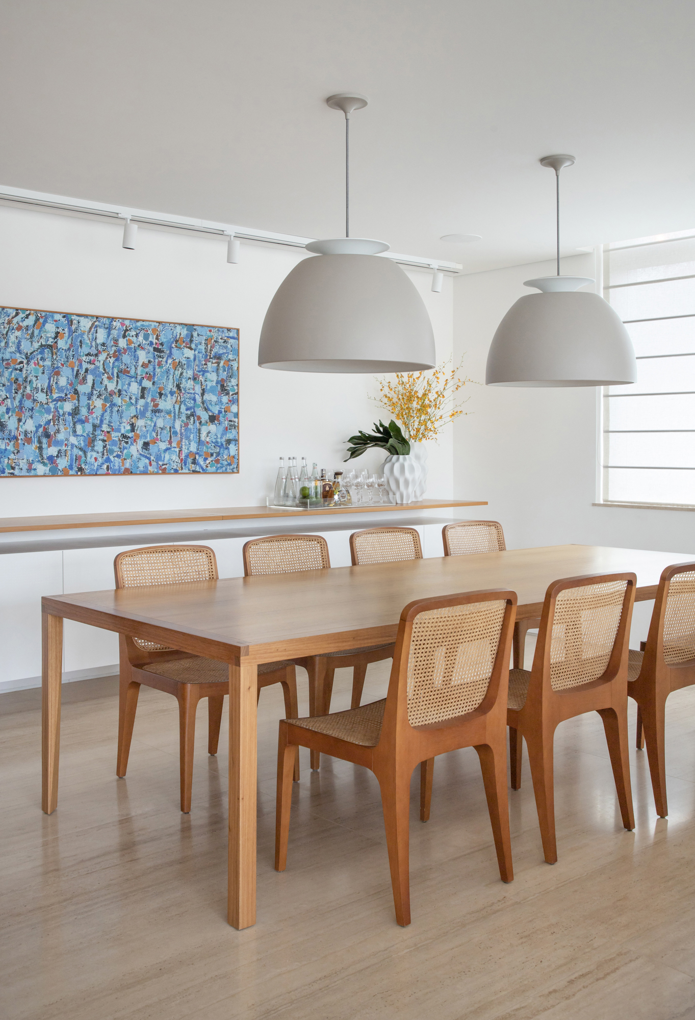 Sala de jantar com mesa de madeira grande, cadeiras de madeira com treliça e luminárias brancas.
