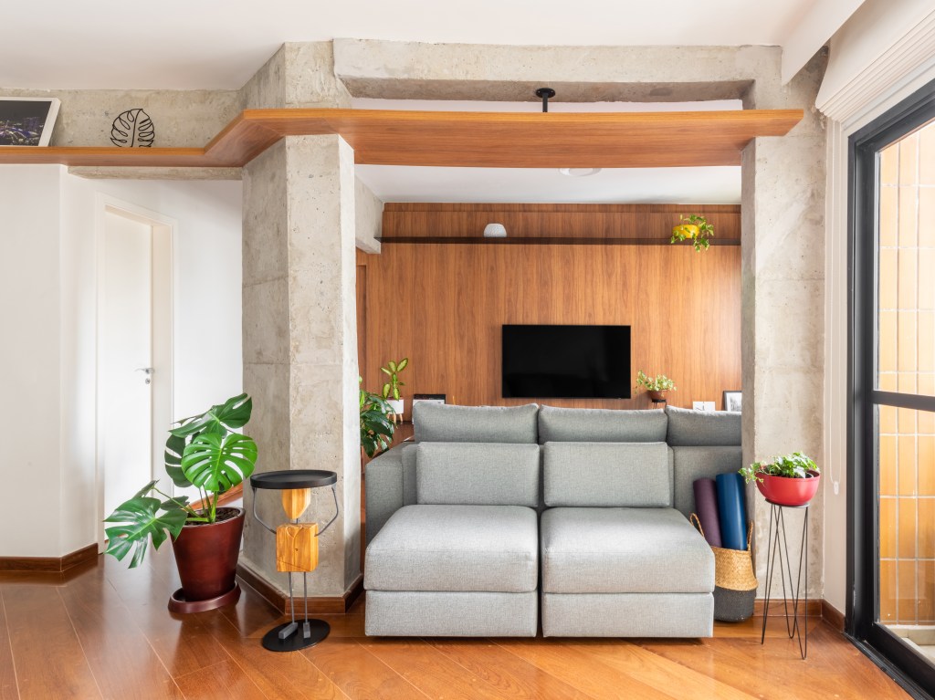 Sala de tv integrada com estar, piso de madeira, sofá cinza e viga de concreto.