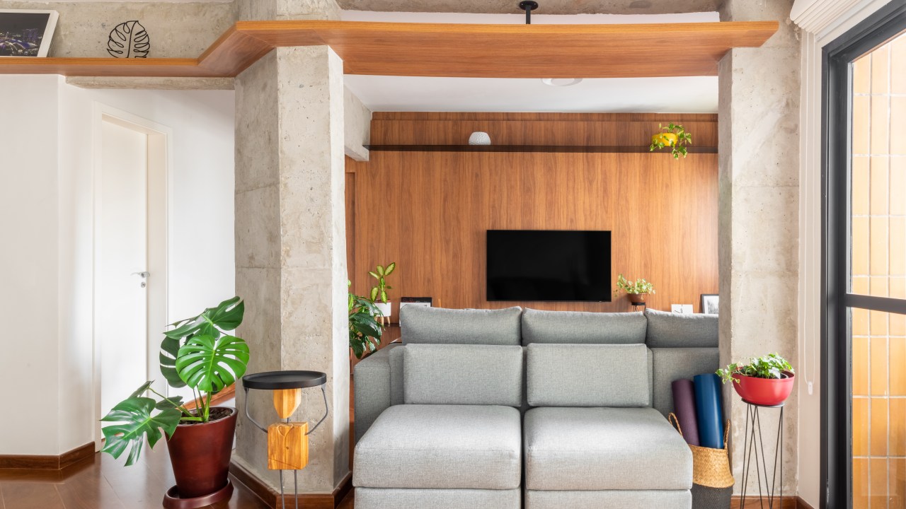 Sala de tv integrada com estar, piso de madeira, sofá cinza e viga de concreto.