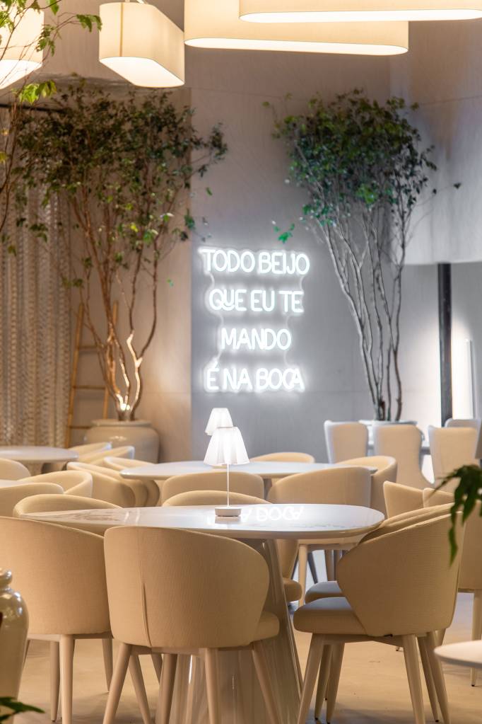 restaurante-grego-sp-inspirado-arquitetura-mykonos-carla-felippi-casacor-sp-credito-favaro-jr (5) Restaurante grego em SP é inspirado na arquitetura de Mykonos. Projeto de Carla Felippi para a CASACOR São Paulo 2023. Na foto, mesas e cadeiras, neon na parede e árvores.