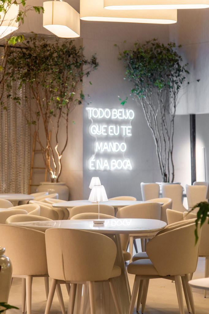 Restaurante grego em SP é inspirado na arquitetura de Mykonos. Projeto de Carla Felippi para a CASACOR São Paulo 2023. Na foto, mesas e cadeiras, neon na parede e árvores.