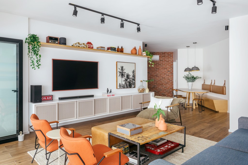 Sala de estar integrada com sofá azul, poltronas laranjas, mesa de centro de madeira, pufe de couro.