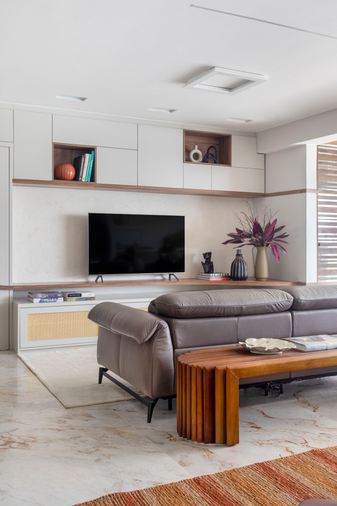 Sala de tv com estante de marcenaria branca, sofá cinza, banco de madeira atrás do sofá.